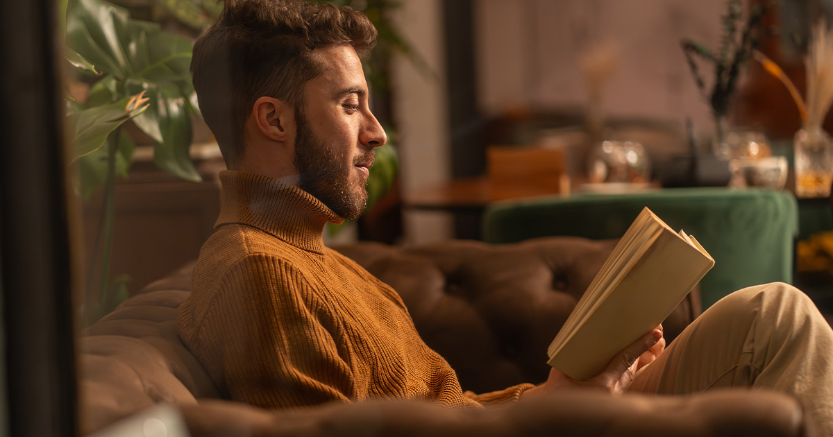 Man sitting on a couch reading a book in a warm, softly lit living room