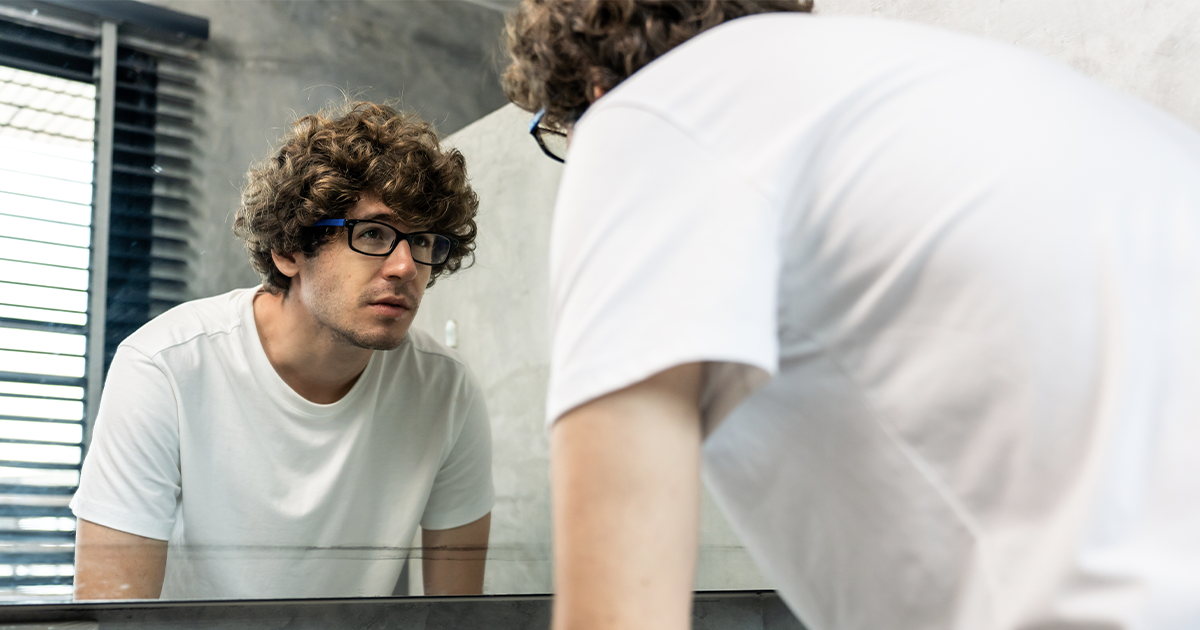 Man wearing glasses and a white shirt looking at himself thoughtfully in the mirror