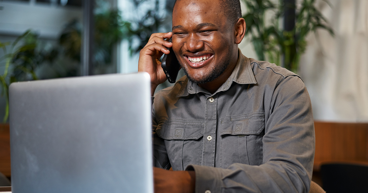 Smiling man talking on the phone while working on a laptop in a bright office setting