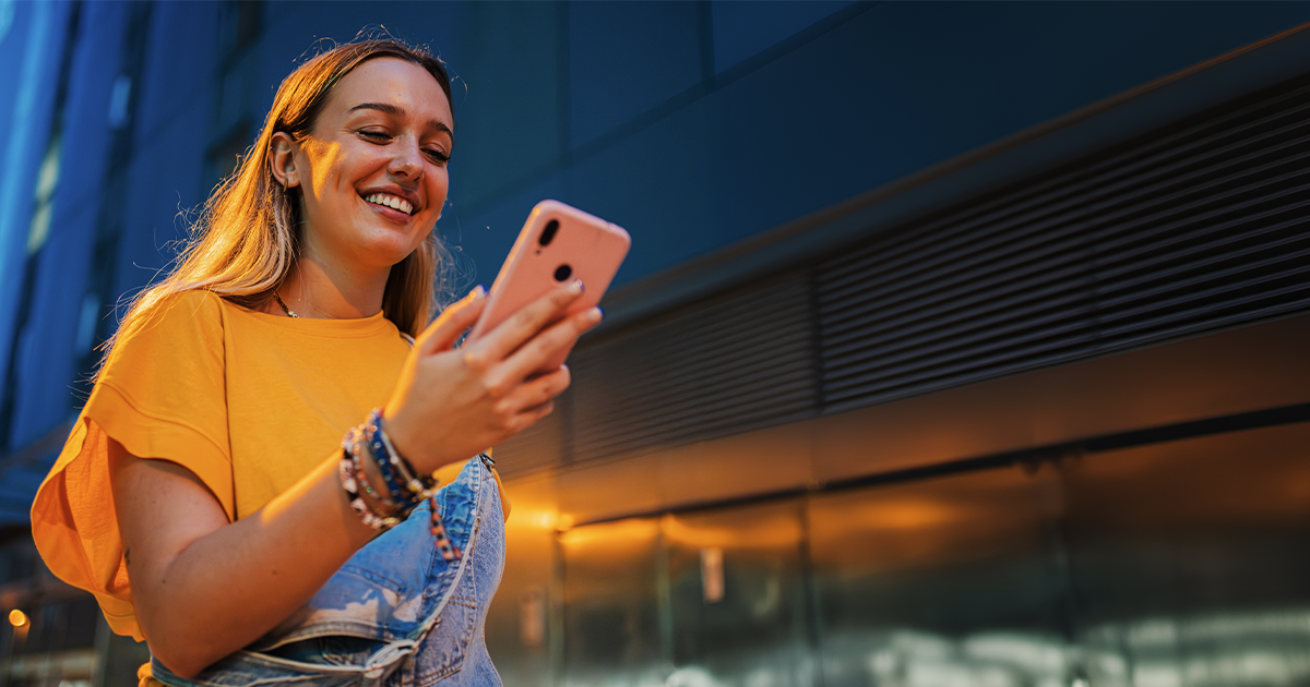 Smiling woman wearing a yellow shirt looking at her smartphone while walking outside in the evening