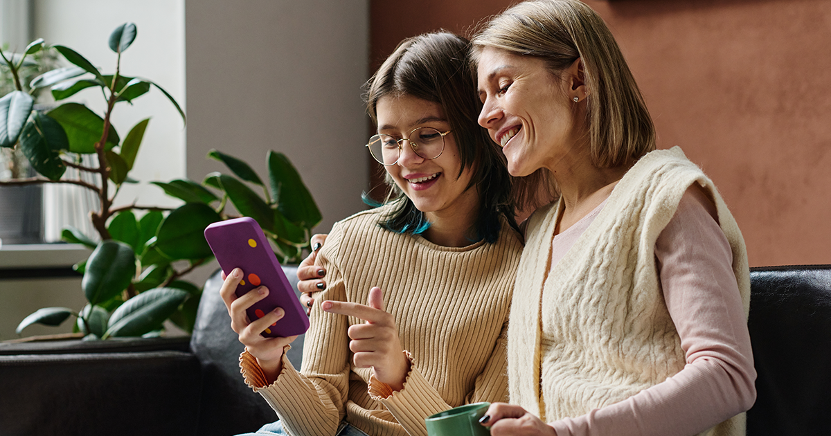 Smiling woman and teenage girl sitting together on a couch looking at a smartphone