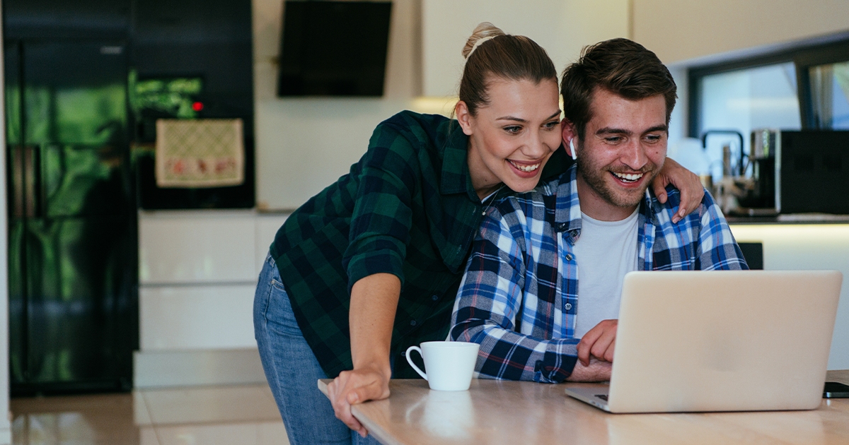 Smiling couple in a kitchen looking at a laptop together while drinking coffee