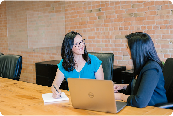 Two women sitting at a conference table talking and smiling, with a laptop and notebook in front of them