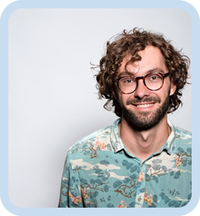 Man with curly hair and glasses smiling while wearing a patterned button-up shirt against a light gray background