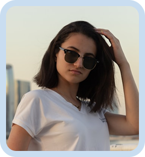 Woman wearing sunglasses and a white shirt brushing her hair back while standing outdoors near city buildings