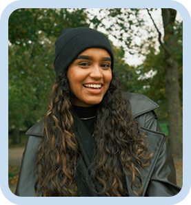 Smiling woman with long curly hair wearing a black beanie and jacket standing outdoors in a park
