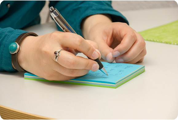 Person writing notes on a blue notepad at a desk while wearing a teal shirt and silver ring