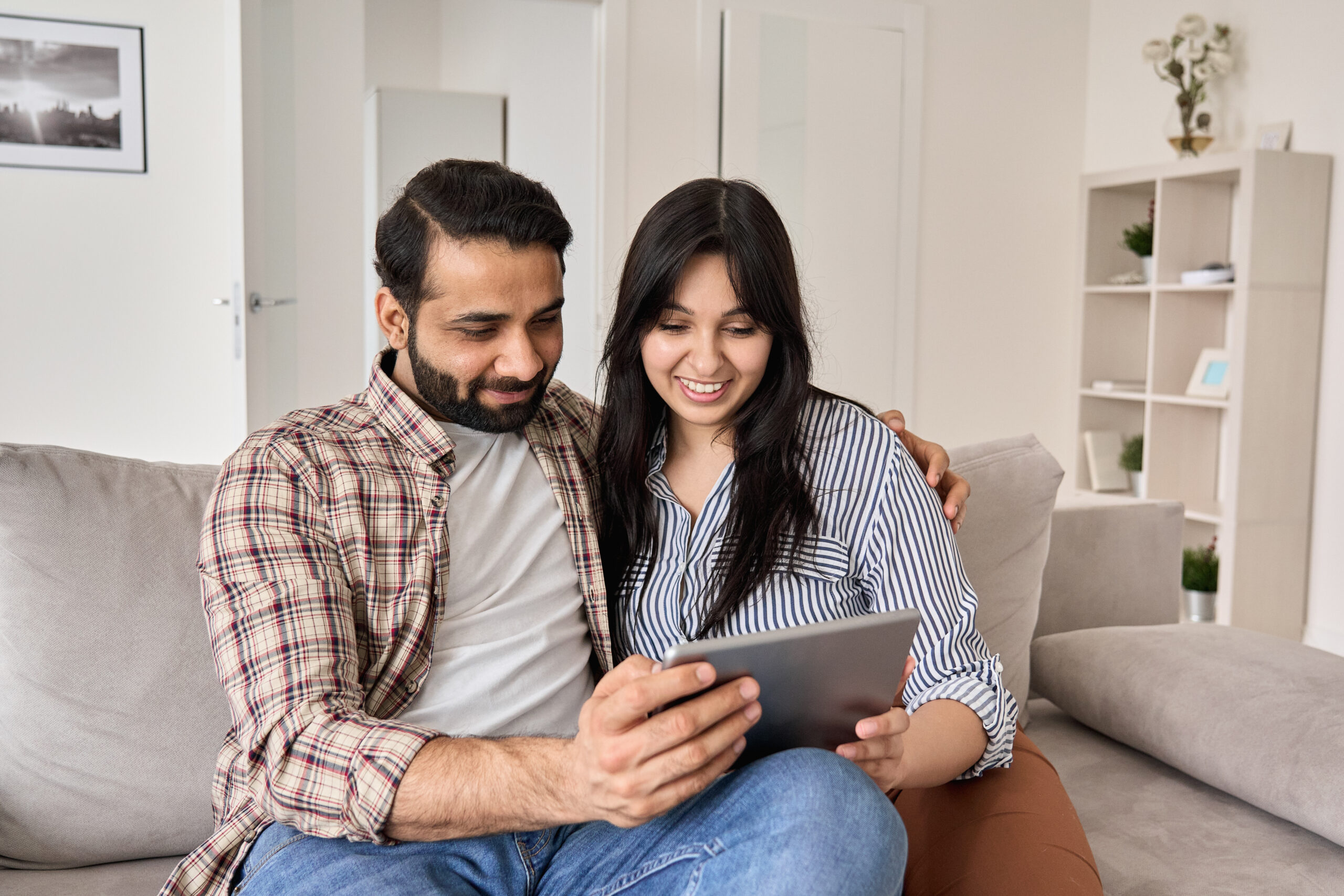 Smiling couple sitting together on a couch looking at a tablet in a bright living room