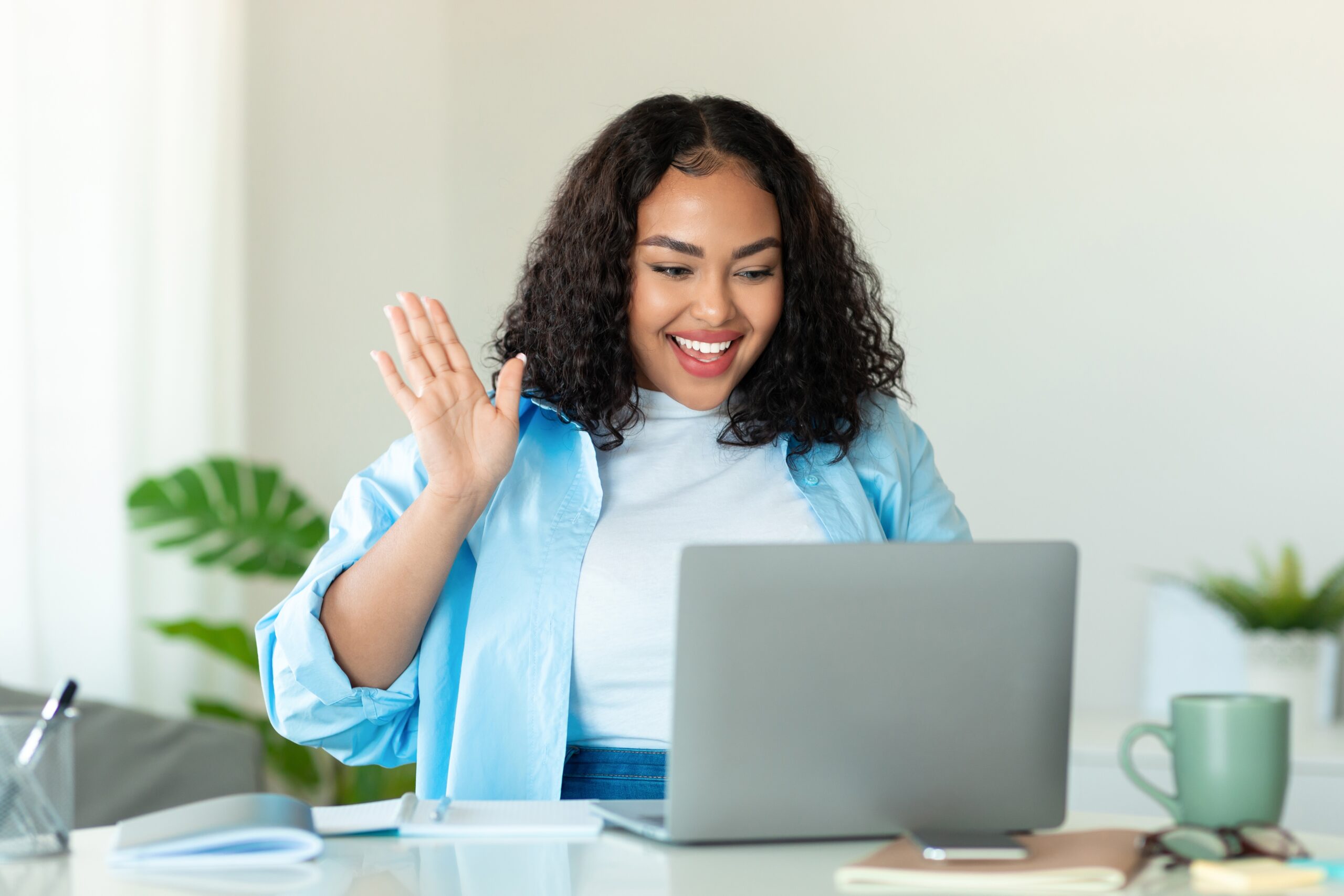 Smiling woman waving during a video call while sitting at a desk with her laptop