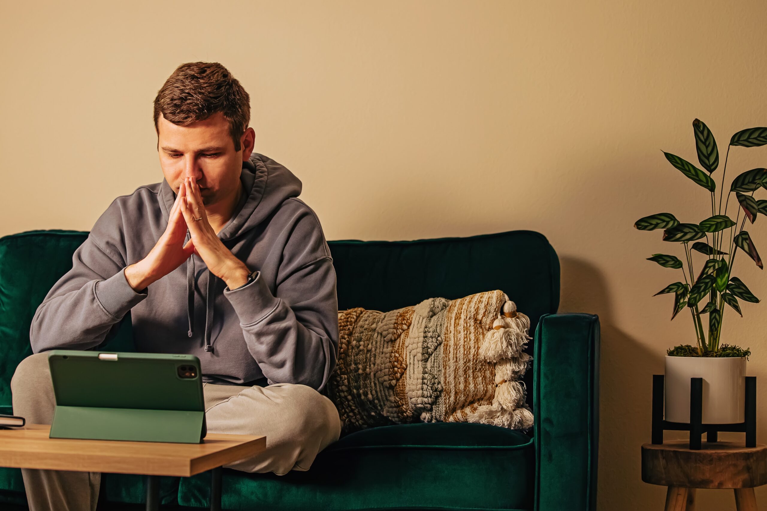Man sitting on a couch looking thoughtful with hands clasped in front of a tablet during a virtual session