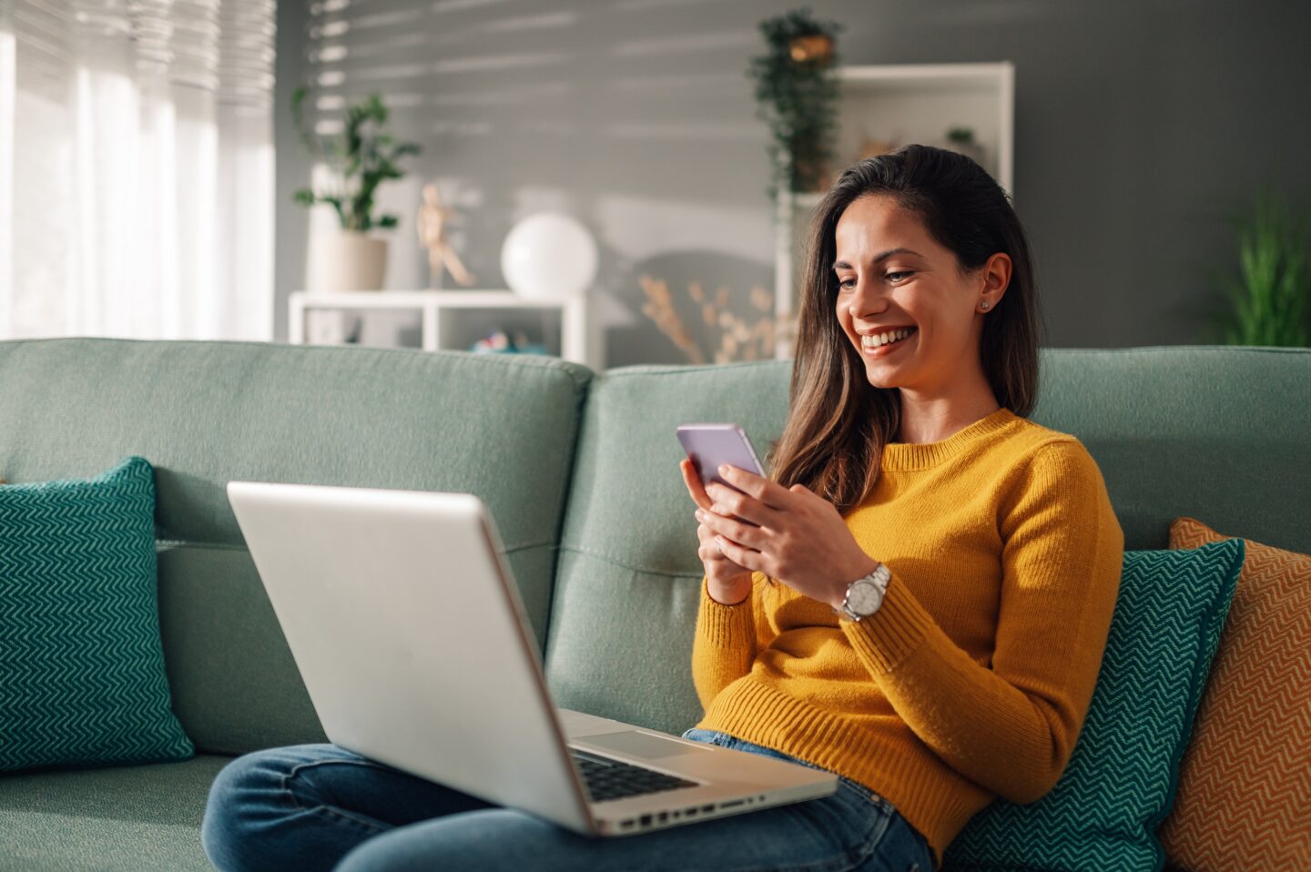 Smiling woman sitting on a couch using her smartphone with a laptop open beside her