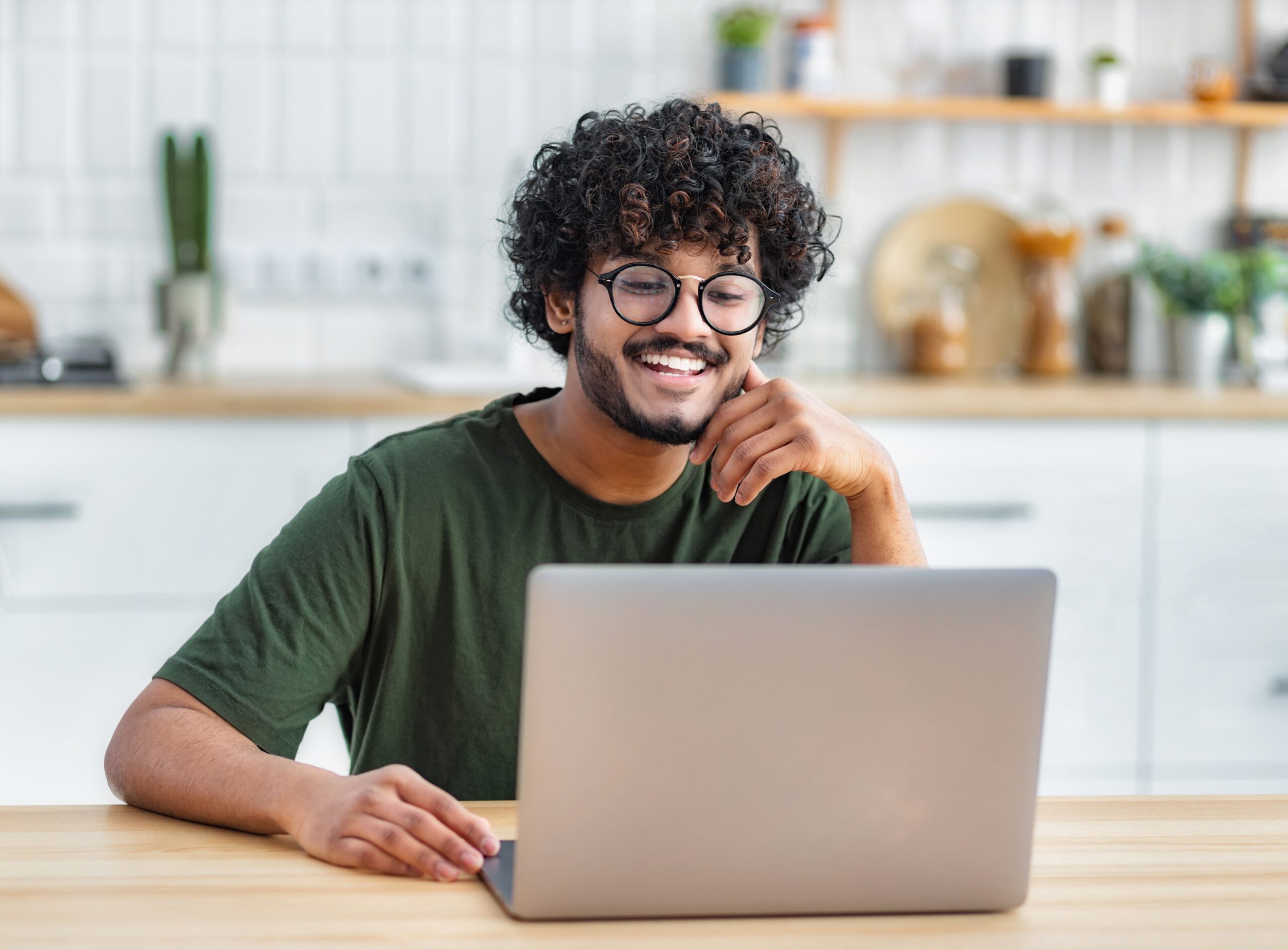 Smiling young man with curly hair and glasses working on a laptop in a bright kitchen