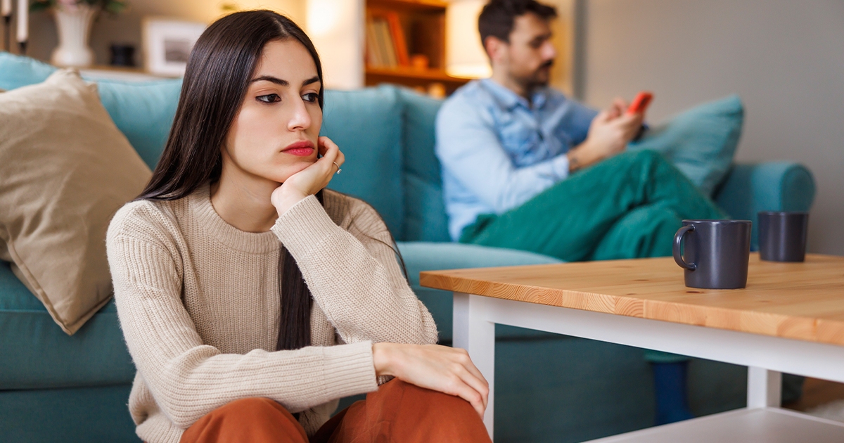 Woman sitting on the floor looking upset while a man sits in the background using his phone