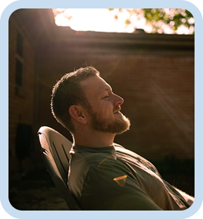 Man with short hair and beard relaxing outdoors in a chair, smiling in the sunlight near a brick building