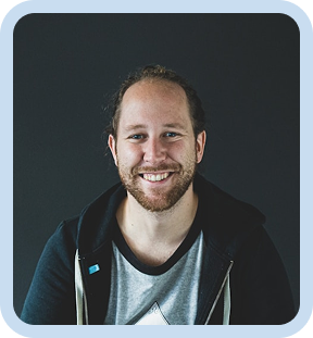 Smiling man with beard and long hair tied back wearing a hoodie and t-shirt against a dark background